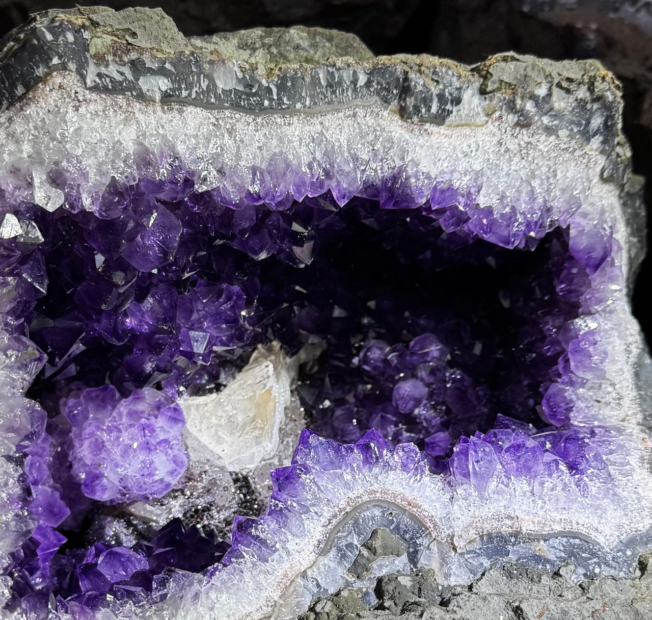 Geoda de amatista abierta, vista en corte: costra de roca gris por fuera, una banda de ágata gris azulada, cuarzo blanco, amatista violeta hacia el hueco y un cristal blanco de calcita
