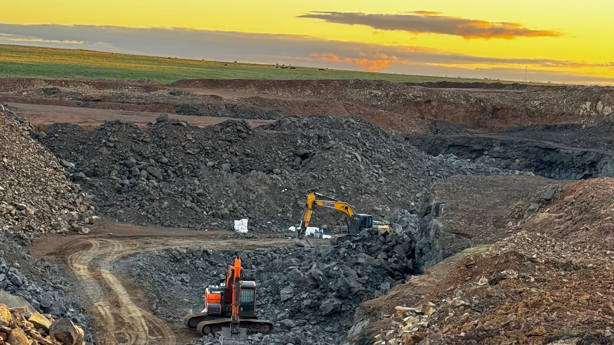Pozo de una cantera de Los Catalanes al atardecer: una excavadora amarilla y otra naranja trabajando entre pilas de roca, con bolsones blancos y el campo en el horizonte